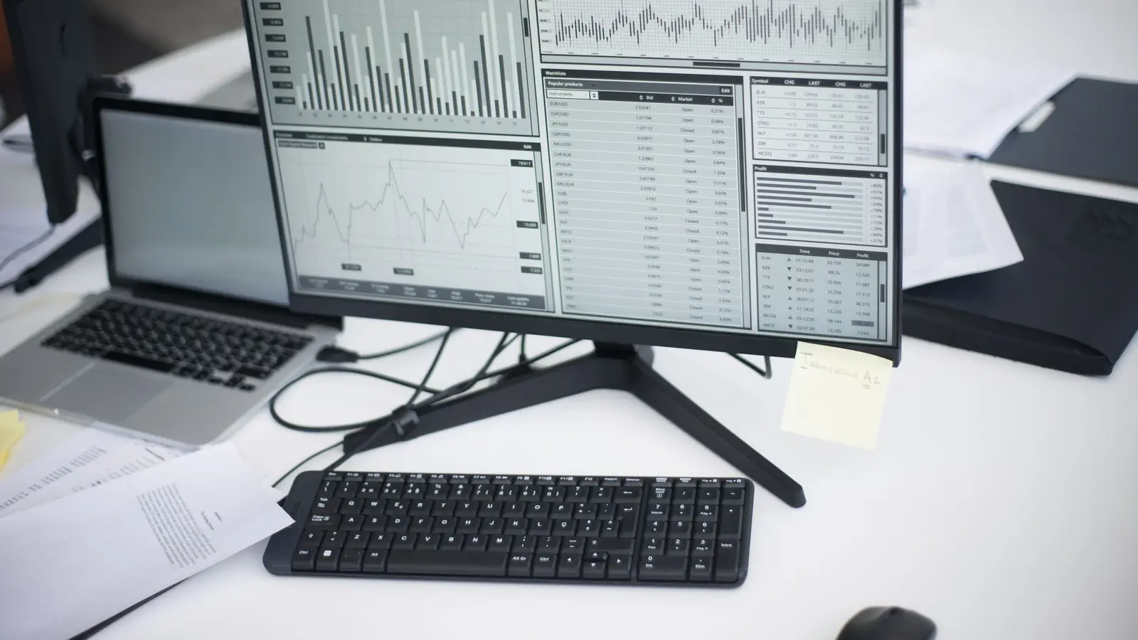 A desk monitor showing charts and data tables beside a laptop and printed reports