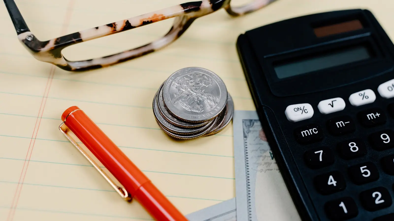 A calculator, a red pen, coins and reading glasses resting on a notepad of figures