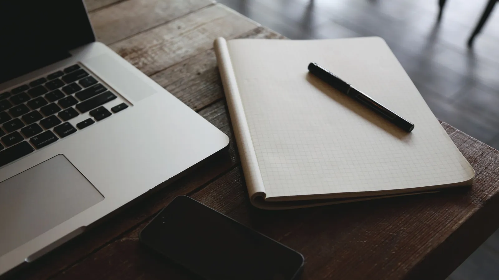 An open laptop beside a squared notebook, a black pen and a phone on a wooden desk