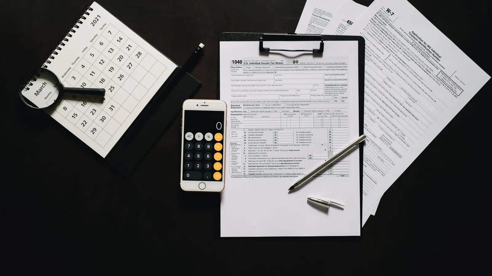 US tax forms on a clipboard beside a phone calculator, a desk calendar and a magnifying glass
